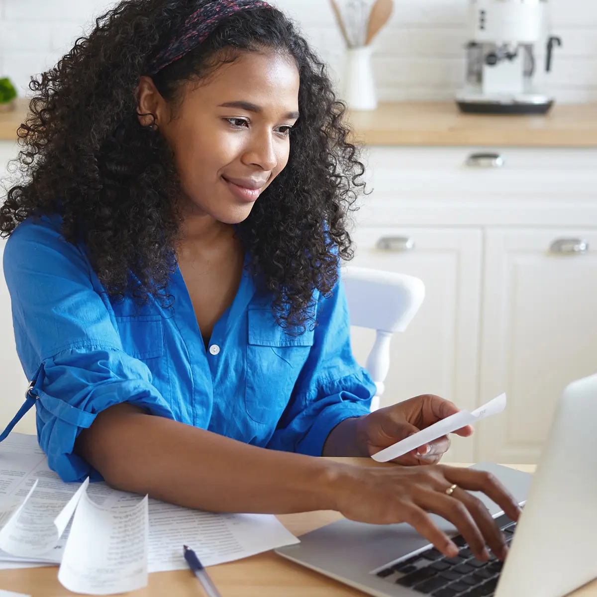 Candid shot of focused beautiful young African housewife wearing headband and casual shirt paying utility bills online using laptop computer, sitting at kitchen table with papers and smiling Man sitting on bench outside.